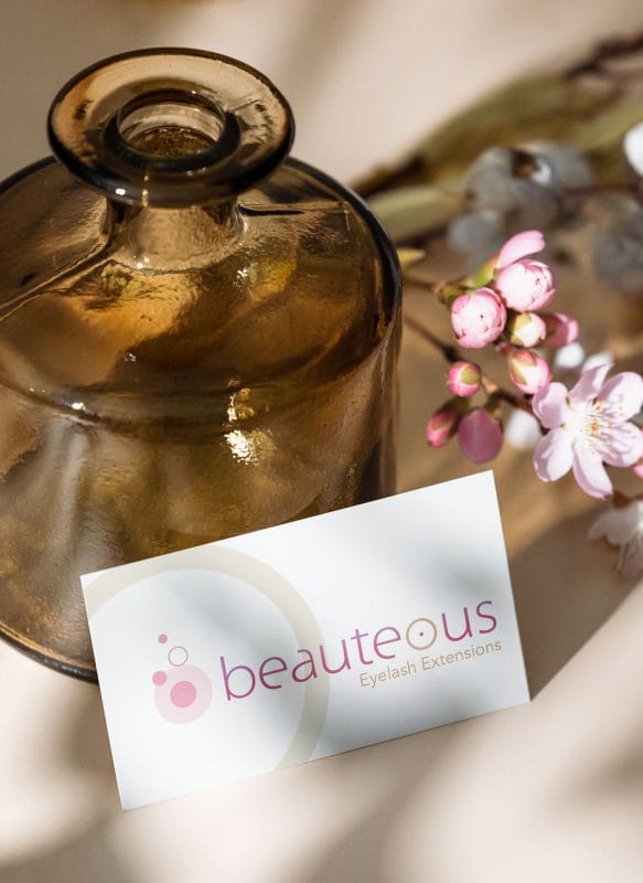 Mockup of Beauteous eyelash extension salon business card placed beside glass bottle and flowers in a styled lifestyle setting.
