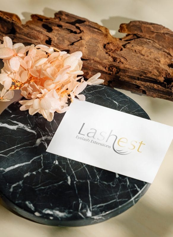 Lashest eyelash extensions business card displayed on a marble surface with soft floral arrangement and natural wood accents.